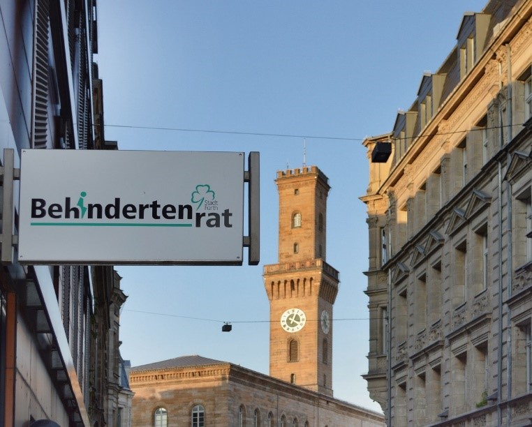 m Vordergrund ragt auf der linken Seite das Schild des Behindertenrats über dem Eingang der Hirschenstraße 2a ins Bild. Mittig steht der Fürther Rathausturm in der späten Nachmittagssonne.