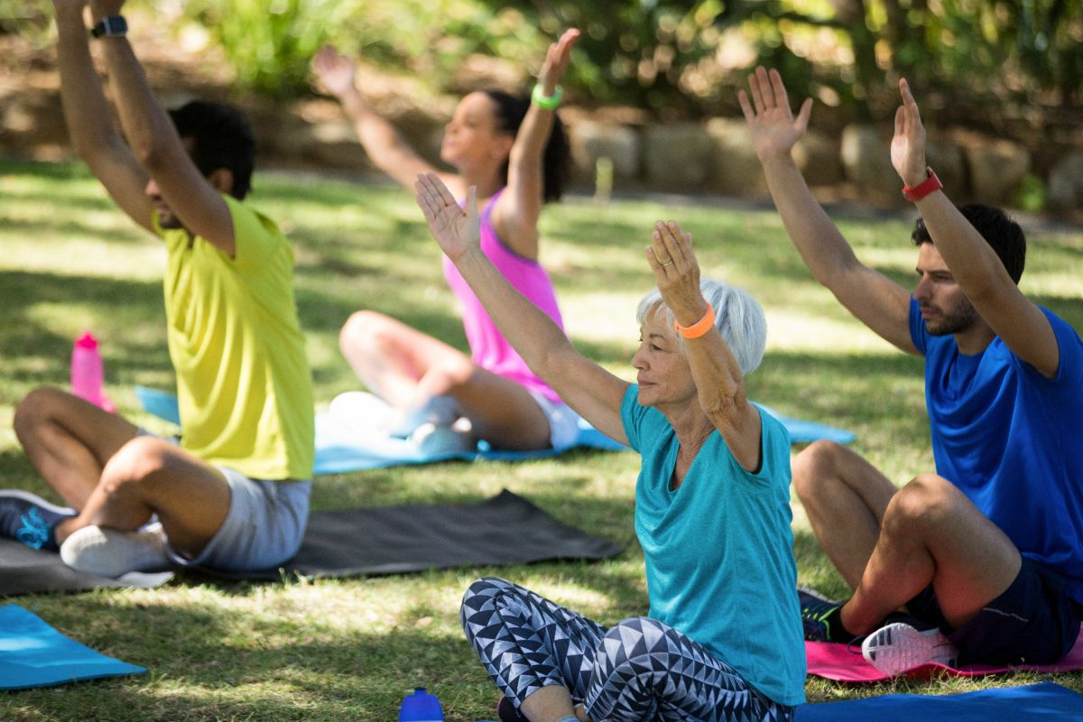 Man sieht eine Gruppe von Menschen unterschiedlichen Alters beim Yoga im Park, gezeigt wird eine Übung im Schneidersitz mit nach oben ausgestreckten Armen.