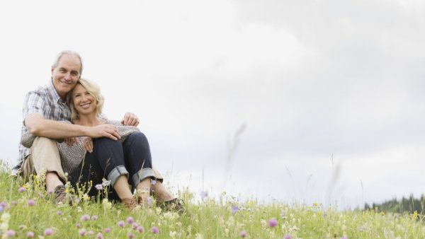 Ein älterer Mann und eine Frau sitzen lächelnd aneinander gelehnt auf dem Boden in einer Blumenwiese. Bildnachweis: Adobe Stock